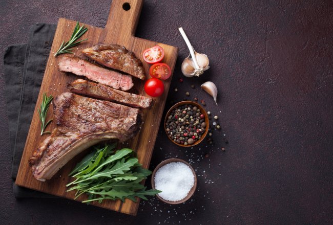 Grilled cowboy beef steak, herbs and spices on a dark stone background. Top view with copy space for your text